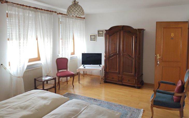 Schrank, TV auf Tischchen, Stuhl mit rundem Beistelltisch, 2 Fenster, Sessel mit blauem Bezug. 